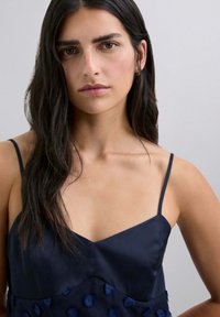 Marinblå satinklänning med tunna remmar, med en v-ringning och texturerad blommönsterapplikation vid midjan. Slät tyg med subtil lyster.