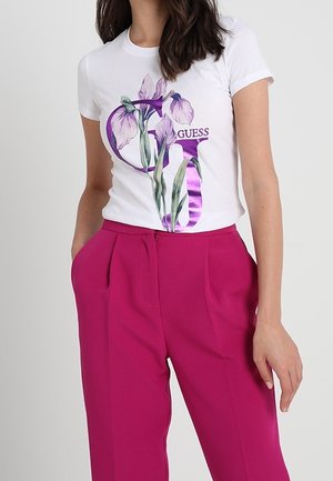 Femme portant un t-shirt blanc avec des motifs floraux violets et le logo "GUESS", associé à un pantalon taille haute magenta vif, les mains dans les poches.