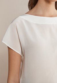 Lekka bluzka w kremowym kolorze z dekoltem łódką, krótkimi rękawami oraz gładką, satynową teksturą o subtelnym połysku.