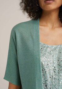 Zelený pletený kardigan s krátkými rukávy, přesáklý flitrovaným topem se stříbrnými a zelenými flitry, hladká textura a zaoblený výstřih.