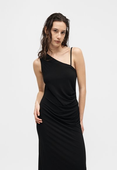 Νέα γυναίκα με βρεγμένα, κυματιστά μαλλιά που φορά στενή μαύρη φόρεμα με έναν ώμο και ρυτιδωμένη λεπτομέρεια, στέκεται μπροστά σε ένα απλό λευκό φόντο.