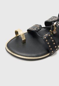 Sandal nero con dettagli dorati, cinturini chiodati e un anello per le dita. Realizzato in materiale liscio con suola piatta e bordo metallico.