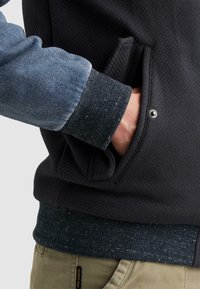 Mano en el bolsillo lateral de una chaqueta negra texturizada con botón a presión, vistiendo manga azul y pantalones beige con etiqueta negra.