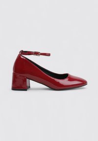 Chaussure basse en cuir verni rouge avec un talon carré, bout pointu et une bride de cheville avec une petite boucle en or, présentée en profil latéral.