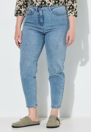 Person trägt hellblaue High-Waist-Jeans und grüne Wildleder-Slipper und steht an einer weißen Wand.