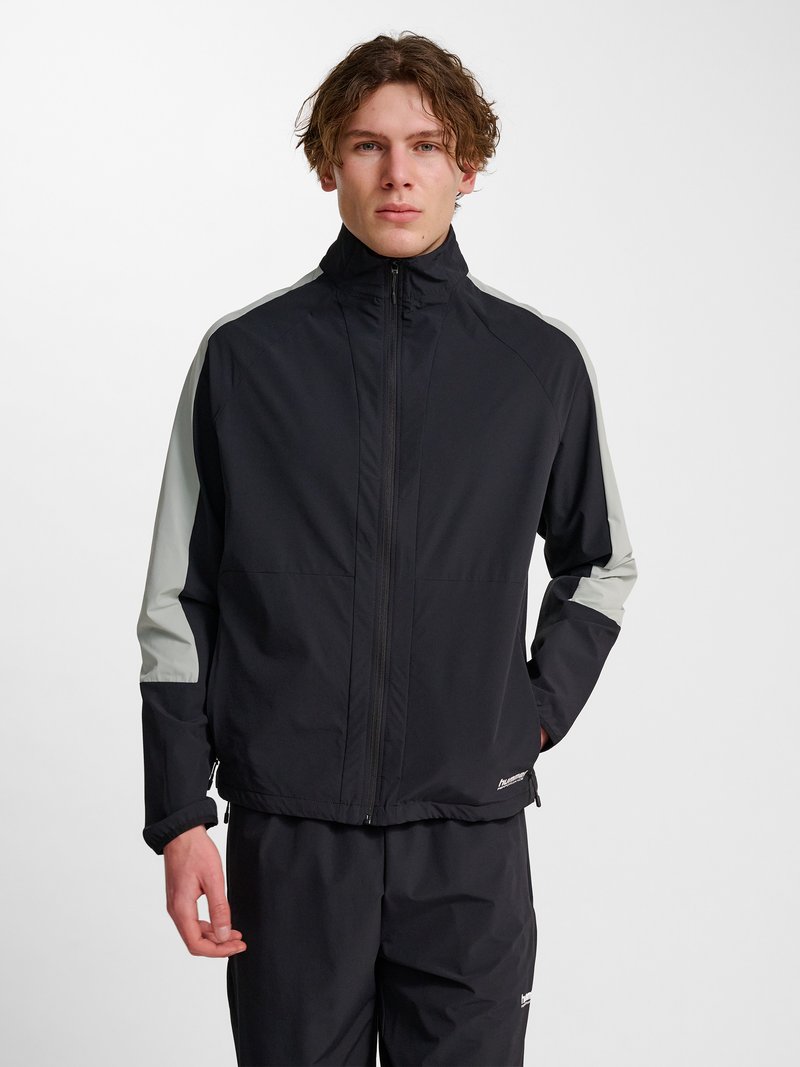 Jonge man met kort krullend haar draagt een zwart jack met rits en grijze panelen op de mouwen en bijpassende zwarte broek tegen een witte achtergrond.