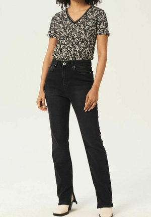 Kobieta ubrana w czarną kwiecistą bluzkę z krótkim rękawem i dekoltem w serek, czarne jeansy z wysokim stanem i rozcięciami po bokach oraz jasne botki na obcasie.