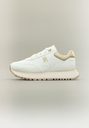 Zapatilla blanca y beige con detalles dorados, vista lateral, con cordones, logotipo T-H y suela texturizada sobre un fondo neutro.