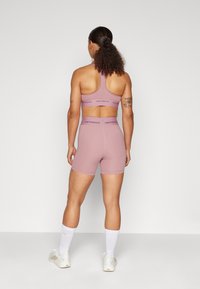 Mulher com cabelo encaracolado vestindo um top desportivo mauve da New Balance e calções a condizer, com meias brancas e sapatilhas bejes, em pé contra um fundo simples.