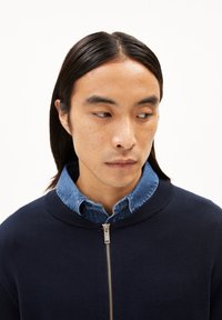 Marineblauwe zip-up sweater met geribde kraag en textuur, gedragen over een lichtdenim overhemd. Voorzien van een zilveren rits en een minimalistisch ontwerp.