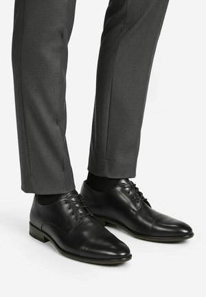 Schwarze, klassische Herrenschuhe aus Leder mit Schnürung, getragen zu dunkelgrauen Anzughosen und schwarzen Socken, stehend auf einer weißen Oberfläche.