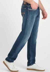 Jeans de mezclilla azul con un corte recto, textura desgastada y un ligero efecto de desgaste. Cuenta con cinco bolsillos y una marca de cuero marrón en la parte trasera.