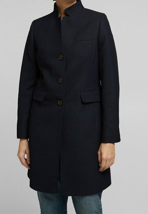 Person trägt einen schwarzen Mantel mittlerer Länge mit drei Knöpfen, zwei Pattentaschen und einem Stehkragen, kombiniert mit blauen Jeans.