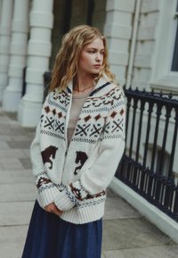 Knit cardigan in white with brown and navy geometric patterns and horse motifs. Features a shawl collar and ribbed cuffs. Worn over a beige top.