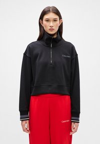 Black zip-up sweatshirt with high neck, featuring Calvin Klein logo in white. Paired with red sweatpants and striped cuffs.