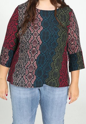 Woman wearing a multicolored snake print blouse with 3/4 pleated sleeves and light blue jeans against a plain background.