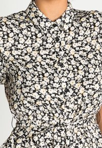 Svart blommig klänning med vita och gula blommor, med krage, knappslutning fram och ett bälte i midjan. Tyget verkar lätt och luftigt.