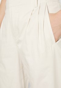 Close-up van beige geplooide broek met een hand gedeeltelijk in de rechterzak, laat de stoftextuur en stikdetails zien.