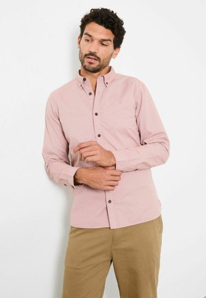 Hellrosa Langarmshirt mit Knopfkragen, dunklen Knöpfen und einem gestickten Logo auf der Brust, kombiniert mit beigen Hosen.