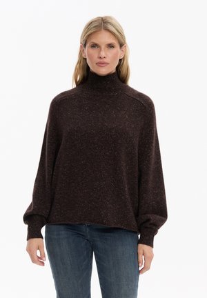 Kobieta o blond włosach, ubrana w ciemnobrązowy sweter z golfem i niebieskie dżinsy, stoi na jednolitym białym tle.