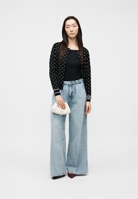 Γυναίκα με μακριά σκοτεινά μαλλιά, που φορά μαύρο πουλόβερ με πουά, μαύρο τοπ και ανοιχτόχρωμα τζιν με φαρδιά σκέλη, κρατώντας μικρή λευκή τσάντα.