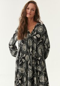 Schwarzes Blumenkleid mit weißem Sonnenblumenmuster, langen Ärmeln, V-Ausschnitt, gerafftem Oberteil und gestuftem Rock für eine lockere Passform. Weicher Stoff.