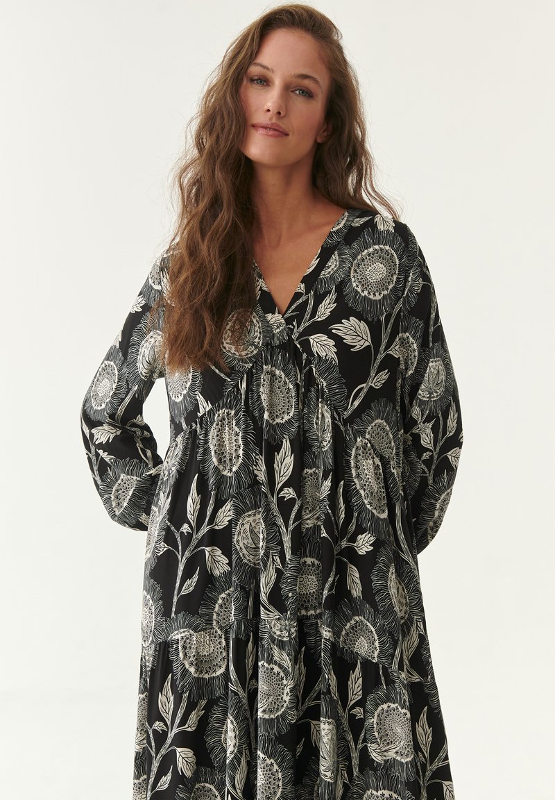 Schwarzes Blumenkleid mit weißem Sonnenblumenmuster, langen Ärmeln, V-Ausschnitt, gerafftem Oberteil und gestuftem Rock für eine lockere Passform. Weicher Stoff.