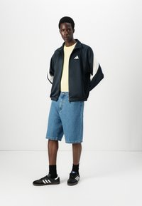 Jacket atlética preta com detalhes brancos, t-shirt amarela, calças de ganga azuis e sapatos pretos com riscas brancas. Tecido suave e estilo casual.