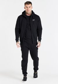 Black zip-up hoodie with a front pocket, paired with black joggers featuring side pockets. Both in a soft, textured fabric. Black sneakers.