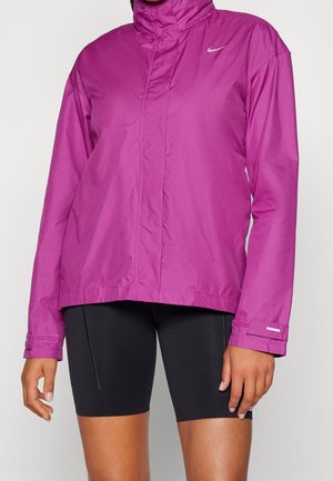 Close-up van een persoon die een felpaarse Nike windbreaker jas en zwarte strakke fietsshort draagt tegen een effen achtergrond.
