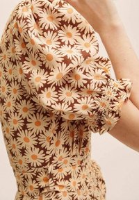Vestido marrón con estampado floral sobre un fondo color crema que presenta margaritas. Mangas abullonadas con fruncido elástico y pequeños puntos centrales naranjas. Tejido suave.