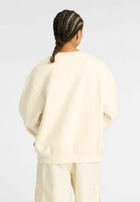 Une personne avec des cheveux tressés se tient de dos, portant un sweat-shirt crème surdimensionné et un pantalon cargo crème, sur un fond uni.