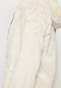 Chaqueta de pelo sintético color crema con capucha, que presenta una superficie texturizada y bolsillos laterales. Cierre de cremallera en la parte delantera.
