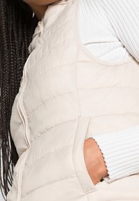 Gilet imbottito beige chiaro con cuciture orizzontali, tasche laterali e chiusura con zip, progettato con una texture liscia e lucida.