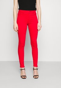 Pantalones vaqueros ajustados rojos con cintura alta y ansas para el cinturón, con cierre de botón y cremallera. Combinados con sandalias negras de tacón alto con tiras.