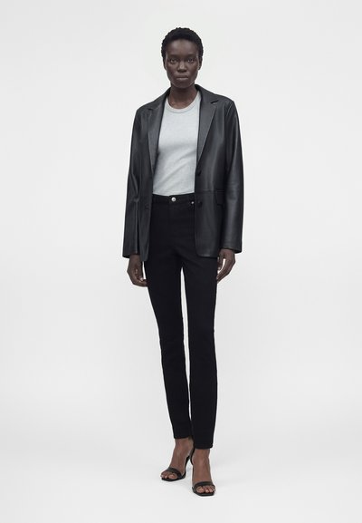 Γυναίκα στέκεται φοράει μαύρο δερμάτινο σακάκι, ανοιχτό γκρι μπλουζάκι με στρογγυλή λαιμόκοψη, μαύρο στενό παντελόνι και μαύρα πέδιλα με ψηλά τακούνια σε απλό λευκό φόντο.
