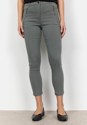 Femme portant un pantalon gris moulant longueur cheville avec des poches avant zippées et des chaussures noires à petit talon, debout sur fond blanc.