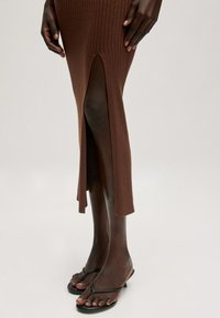 Bruine geribbelde midi-jurk met zijsplitten en een strakke pasvorm, gecombineerd met zwarte platte sandalen, benadrukt textuur en een elegant ontwerp.