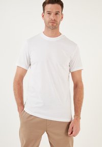 T-shirt bianco in cotone con scollo rotondo e maniche corte, caratterizzato da una texture liscia e una vestibilità comoda. Abbinato a pantaloni beige.