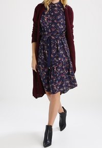 Marineblaues Blumenkleid mit kurzen Ärmeln, taillierter Taille und schwingendem Rock, kombiniert mit einem langen burgunderfarbenen Strickcardigan und schwarzen Stiefeletten.