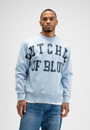 Homme portant un sweat-shirt bleu clair avec un grand texte noir "Dutchie of Blue" et un jean bleu, debout devant un fond clair uni.