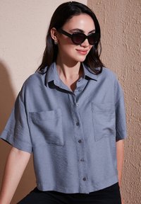 Light blue short-sleeved button-up shirt with two chest pockets, soft textured fabric, and a relaxed fit. Black sunglasses worn for contrast.