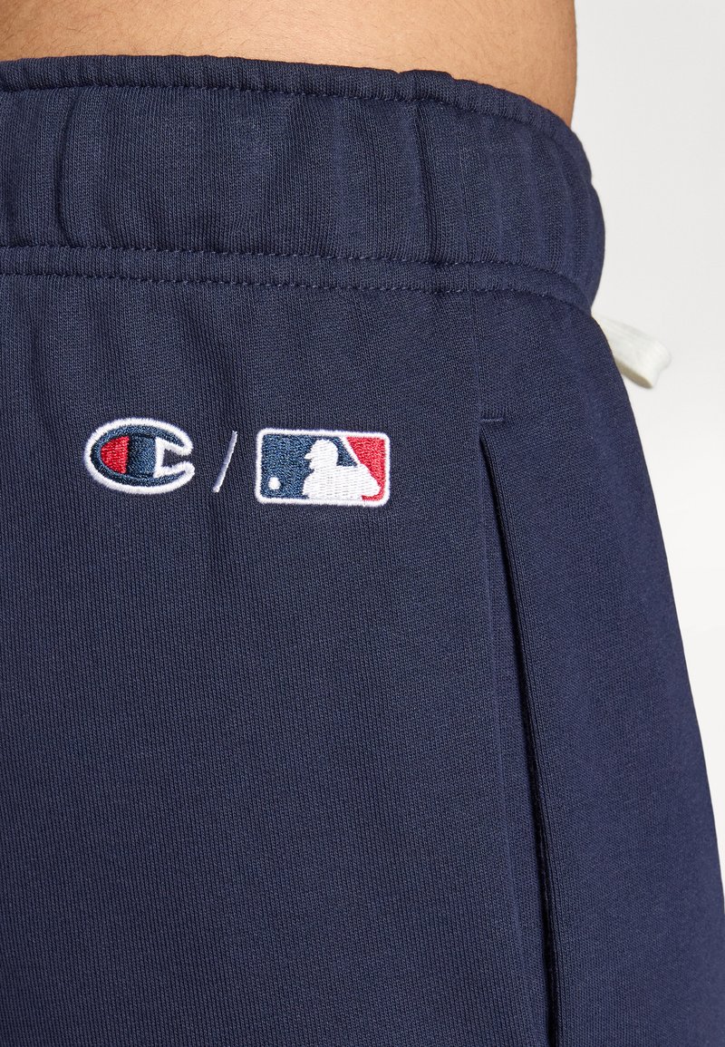 Marineblauwe joggingsbroek met elastische tailleband, voorzien van geborduurd Champion-logo en MLB-logo in rood, wit en blauw. Zachte stoftextuur.