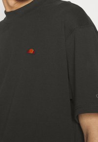 Chemise noire à manches courtes avec un petit logo en demi-cercle rouge et orange sur la poitrine et des coutures visibles sur l'épaule et la manche.