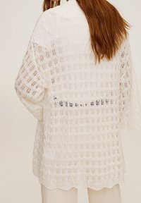 White open-knit cardigan with a loose fit, scalloped hem, and patterned texture. Features wide sleeves and a slightly longer back.