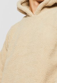Hoodie en polaire beige avec une surface texturée et douce. Caractérisé par une grande capuche et une coupe décontractée, mettant l'accent sur le confort et la chaleur.