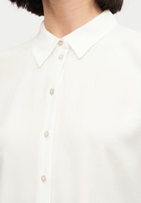 Λευκό πουκάμισο με γιακά και μαργαριταρένια κουμπιά κλειστά μέχρι τον λαιμό, που φοριέται από ένα άτομο με κοντά σκούρα μαλλιά.