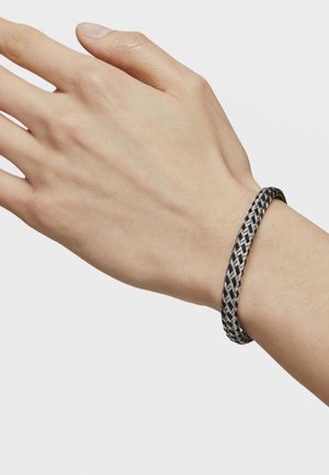 Ręka nosząca czarno-szary pleciony bransoletkę na jednolitym jasnoszarym tle.