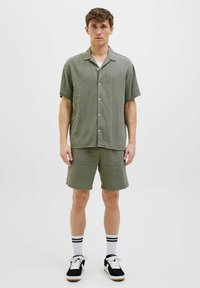 Lyse oliven grøn kortærmet skjorte og matchende shorts lavet af struktureret stof; har knapper og en afslappet pasform. Sorte sneakers med hvide sokker.