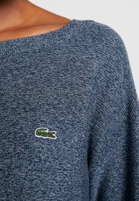 Pull en tricot bleu marine avec un large décolleté et un logo crocodile vert brodé sur le côté gauche de la poitrine. La texture est douce et à motifs.